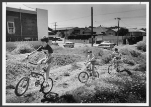 Boys cycling across a vacant section, Karori, Wellington