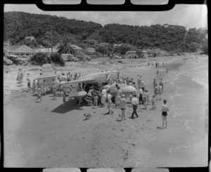 Tourist Air Travel, Grumman Widgeon aircraft ZK-BGQ at Paihia, Northland, shows crowd on the beach and motorcars and houses in the background