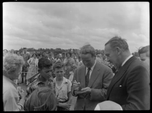 Mr Cummingham and Mr Frank Lloyd standing on runway signing an autograph for unidentified boys and woman with crowd long on, for TEAL Comet aircraft plane, Whenuapai, Auckland