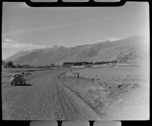 A Morris Minor car on a shingle road at Kingston with Lake Wakatipu, houses and mountains beyond, Central Otago Region