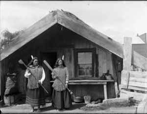 Unidentified Maori women, outside a whare, holding traditional Maori wooden weapons, at Ohinemutu