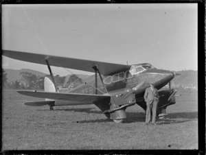 An unidentified man standing alongside a de Havilland DH 90 Dragonfly biplane with 'Air Travel NZ Service' written on side, at an unidentified location