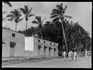Four unidentified RNZAF men walking down a street, near a stone building, Rarotonga, Cook Islands
