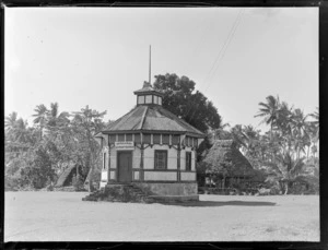 View of the Mau Office hexagonal wooden building with thatch roof huts and trees beyond, Apia, Western Samoa