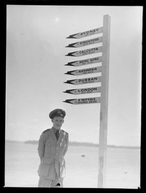 Portrait of Eric Steward Newman, BOAC, next to directional sign, reading from top downwards "To Sydney 1342 Miles, To Singapore 6018 Miles, To Calcutta 8030 Miles, To Hong Kong 8167 Miles, To Alexandria 11980 Miles, To Durban 15548 Miles, To London 14271 Miles, By Empire Flying Boat"