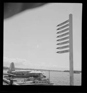 The arrival of the BOAC Hobart G-AGJM, a Short Hythe flying boat, at Mechanic's Bay wharves with motor launches and sign posts showing distances in foreground, Auckland Harbour