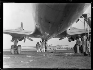 View of the under carriage of PAA's Douglas Clipper DC4 passenger aircraft with unidentified ground personnel loading boxes, [Whenuapai Airfield, Auckland