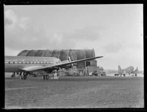 Side view of PAA's Douglas Clipper DC4 passenger aircraft NC 88883 with hangar and plane beyond, [Whenuapai Airfield, Auckland?]