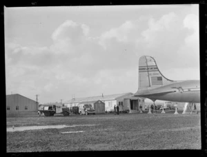 Tail fin view of PAA's Douglas Clipper DC4 passenger aircraft NC 88883 with buildings and vehicles beyond, [Whenuapai Airfield, Auckland?]