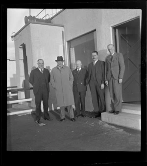 TEA group portrait, Sir Leonard Isitt and F Maurice Clarke with three other unidentified TEA members in front of a building, Mangere Airfield, Auckland