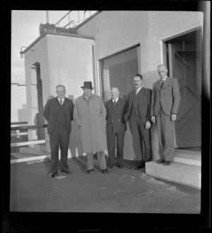 TEA group portrait, Sir Leonard Isitt and F Maurice Clarke with three other unidentified TEA members in front of a building, Mangere Airfield, Auckland