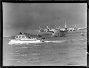 Short Tasman Aircraft (flying boat), Tasman Empire Airways Ltd, showing the Aircraft taxiing towards a boat with an unidentified man standing, with his upper body through a sun roof, Mechanics Bay, Auckland