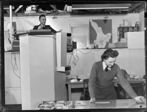 Flying Control Room, Mechanics Bay, Auckland, showing an unidentified man standing on a platform overlooking the unidentified woman working on a chart table