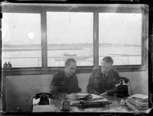 Flying Control Room, Mechanics Bay, Auckland, showing two unidentified men sitting at a desk reading a book, with a seaplane aircraft in the background