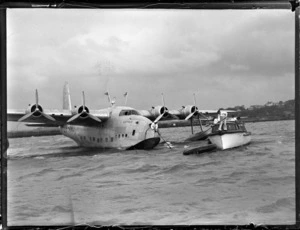 View of the TEAL Short Tasman Flying Boat ZK-AMD 'Australia' being hitched to a mooring with the help of unidentified crew on a launch, Mechanic's Bay, Auckland City