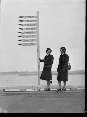 Portrait of Miss B P Morton and Miss L M Magnus, TEAL Stewardesses in uniform with wharf signpost, Mechanic's Bay, Auckland City