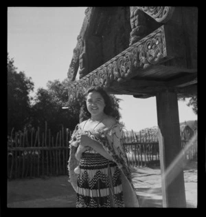Unidentified Maori girl standing under carved Pataka, wearing traditional Maori dress, bodice with taniko design, piupu and Korowai cloak holding a poi, Whakarewarewa, Rotorua