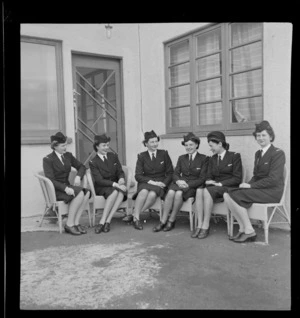 Tasman Empire Airways Ltd stewardesses, (l to r): Misses [Maynes?], Everand, Beckett, Woolley, Paterson and [Martin?] sitting on cane chairs in front of the door of the TEAL building, Mechanics Bay, Auckland