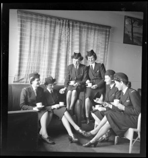 Tasman Empire Airways Ltd stewardesses, (l to r): Misses [Maynes?], Everand, Woolley, [Martin?] and Paterson sitting on cane chairs and a leather couch drinking cups of tea, in the lounge room at the TEAL building, Mechanics Bay, Auckland