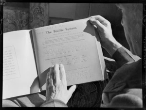 View of an unidentified man holding a Blind Institute 'How to Learn to Read - The Braille System' tutor book, [Auckland?]