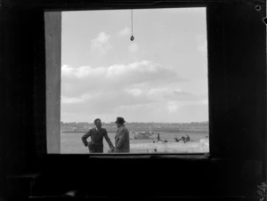 View through the TEAL waiting room window to two unidentified men with the Short Tasman flying boat 'New Zealand' beyond, Mechanic's Bay, Auckland City