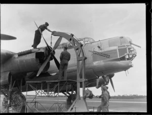 A group of unidentified men using a mobile crane at Whenuapai Airbase, Auckland, preparing to lift the propellor off the Avro Lincoln bomber aircraft Excalibur, showing one of the men standing on the mobile crane and another man standing on the aircraft engine holding onto a propellor blade