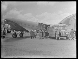 New Zealand National Airways Corporation (NZNAC) Douglas Dakota aeroplane at RNZAF Station, Whenuapai, Auckland, showing unidentified passengers disembarking and a Johnston's Airways Transport bus