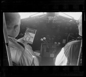 Inside a Dakota transport plane's cockpit with unidentified pilots looking at 'Fighters' book written by Leo White of Whites Aviation, location unknown
