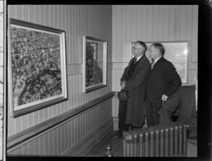 Two unidentified men look at large aerial photographs on a wall, at an exhibition