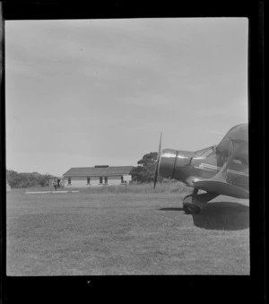Partial view of a bi-plane on a grass runway with unidentified men beyond, Onerahi Aerodrome, Whangarei, Northland region