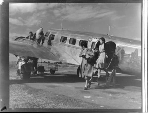 Two unidentified female passengers disembarking NAC Lockheed Electra aeroplane 'Kaka' ZK-AGK, as aircraft engineers work on engine, at an unidentified location, probably Auckland