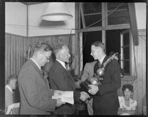 Unidentified men at the RNZAC (Royal New Zealand Aero Club) Pageant event, one man presenting the Herald Cup to another man, the Wigram Cup is in the background