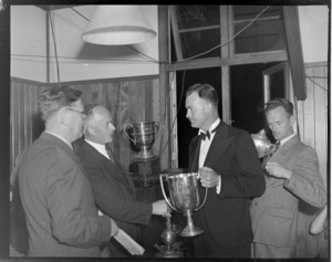 Unidentified men at the RNZAC (Royal New Zealand Aero Club) Pageant event, one man presenting the Cory Wright Cup to another man, the Wigram Cup sits in the background