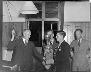 Unidentified men at the RNZAC (Royal New Zealand Aero Club) Pageant event, one man presenting the Bledisloe Cup to another man, while he holds a microphone hanging from the ceiling