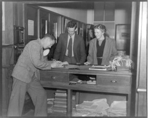 Unidentified man and woman, booking tickets with an unidentified NZNAC (New Zealand National Airways Corporation) booking agent at a Wellington office