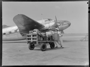 Two unidentified members of New Zealand National Airways Corporation ground crew pushing a trolley loaded with luggage, showing Lochkheed Lodestar aeroplane 'Kotare' ZK-AJM, in background, Rongotai Airport, Wellington