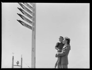 Tasman Empire Airways Ltd stewardess Miss Magnuss, holding a small unidentified boy, next to signpost [at a photoshoot for an advertsing campaign?], Mechanics Bay, Auckland