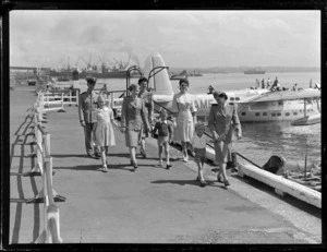 Tasman Empire Airways Ltd crew and passengers, [models for an advertising campaign?] all unidentified, on wharf with Short Sandringham flying boat ZK-AME docked in background, Mechanics Bay, Auckland