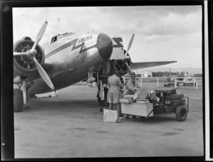 Two unidentified men collecting luggage from a trolley alongside New Zealand National Airways Corporation Lockheed Lodestar aeroplane 'Kotuku', at Palmerston North Airport