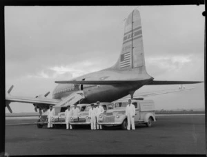 Portrait of Pan American World Airways (PAWA) staff members standing alongside a line-up of trucks, in front of a Douglas Clipper DC-4 passenger aircraft (NC 88883), [Whenuapai Airbase Auckland?]