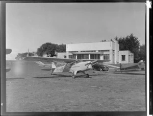 Auster ZK-AOB aircraft, outside a building, including another aircraft and motorcar, Mangere airport, Auckland