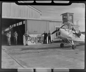 View of D Grieg in centre with unidentified men in front of Aircraft Services Ltd plane hangar and a damaged Auckland Aero Club Tiger Moth aeroplane, Mangere Airfield, Auckland City