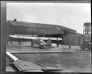 View of Auster J-1B Autocrat ZK-AOB passenger plane with unidentified men in front of a large wooden building, Blenheim [Airport?], Marlborough Region