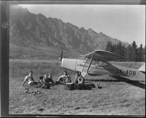 View of unidentified people sitting on grass in front of Auster J-1B Autocrat ZK-AOB passenger plane with the Remarkable Mountain Range behind, Queenstown, Otago Region