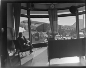 Interior view of an unidentified man, looking out through glass windows at passengers disembarking a NZNAC (New Zealand National Airways Corporation) Dakota ZK-AOJ aircraft, Harewood airport, Christchurch