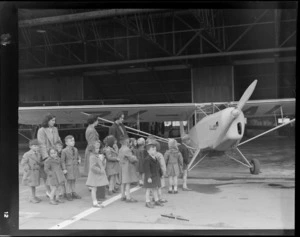 Young school children, standing by an Auster airplane, accompanied by three unidentified women, Rongotai airport, Wellington