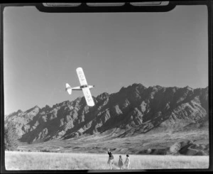 Auster Autocrat monoplane, ZK-AOB, flying low near The Remarkables, Queenstown, as three unidentified children wave to the aircraft from ground