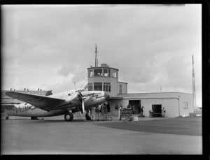 Aircraft Lockhead Lodestar 'Kotare' ZM-AJM, including control tower and passengers, with a man loading baggage into cargo hold of aircraft, at Harewood Aerodrome (Christchurch International Airport)