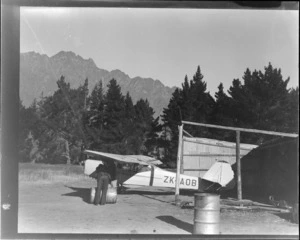 An unidentified man with ZK-AOB Auster J1B Aiglet aeroplane, outside a corrugated-iron hangar, Queenstown, Otago region
