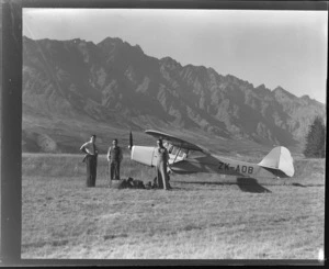 Mr Leo Lemuel White with two unidentified men, standing next to aircraft ZK-AOB Auster J1B Aiglet, including baggage and camera on ground, Queenstown, Otago region